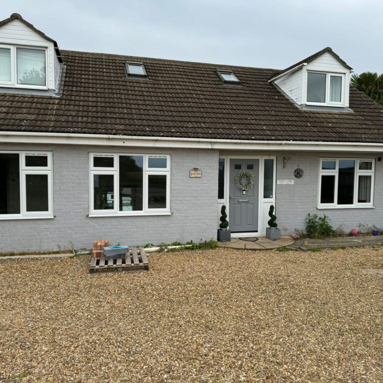 A grey house with a gravel driveway and small garden, featuring a front door and windows.