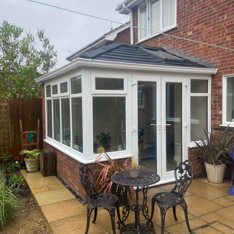 Conservatory with large windows, a table and chairs, surrounded by plants.